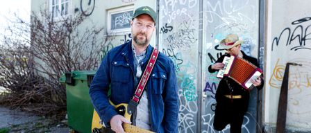 Zwei Männer stehen vor einer mit Graffiti bedeckten Wand. Ein Mann hält eine Gitarre und der andere spielt Akkordeon.