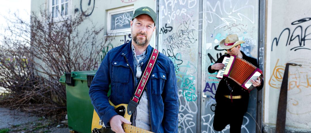 Zwei Männer stehen vor einer mit Graffiti bedeckten Wand. Ein Mann hält eine Gitarre und der andere spielt Akkordeon.