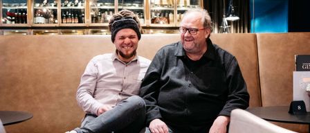 Zwei Männer sitzen auf einer Couch in einem Loungebereich und lächeln für ein Foto. Einer trägt ein rosa Hemd und Jeans, der andere ein schwarzes Hemd und Lederhosen. Sie sind umgeben von moderner Einrichtung und einem Regal mit Flaschen.