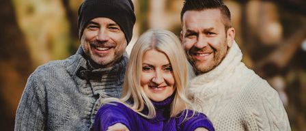 Drei Menschen, zwei Männer und eine Frau, stehen draußen in einem Wald. Sie alle lächeln und scheinen für ein Foto zu posieren.