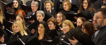 Ein Chor aus Männern und Frauen in Schwarz singt gemeinsam und hält Notenblätter in den Händen, während sie in einer Kirche stehen.