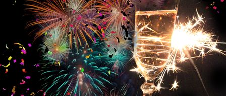 Eine Person hält ein Weinglas mit einem Funkenfeuerzeug im Inneren, umgeben von bunten Feuerwerken und Konfetti am Nachthimmel.