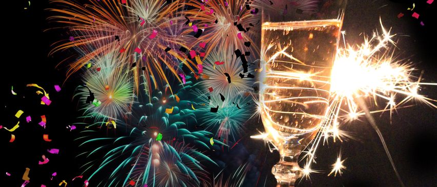 Eine Person hält ein Weinglas mit einem Funkenfeuerzeug im Inneren, umgeben von bunten Feuerwerken und Konfetti am Nachthimmel.