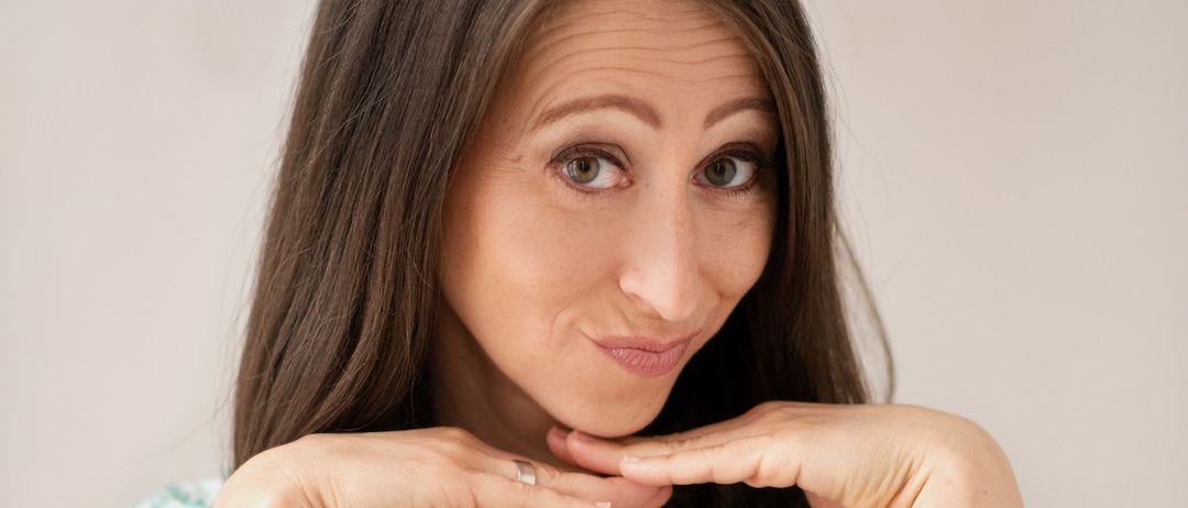 Eine Frau mit braunen Haaren in einem Blumenkleid posiert, berührt ihr Kinn mit beiden Händen und scheint ein lustiges Gesicht zu machen.