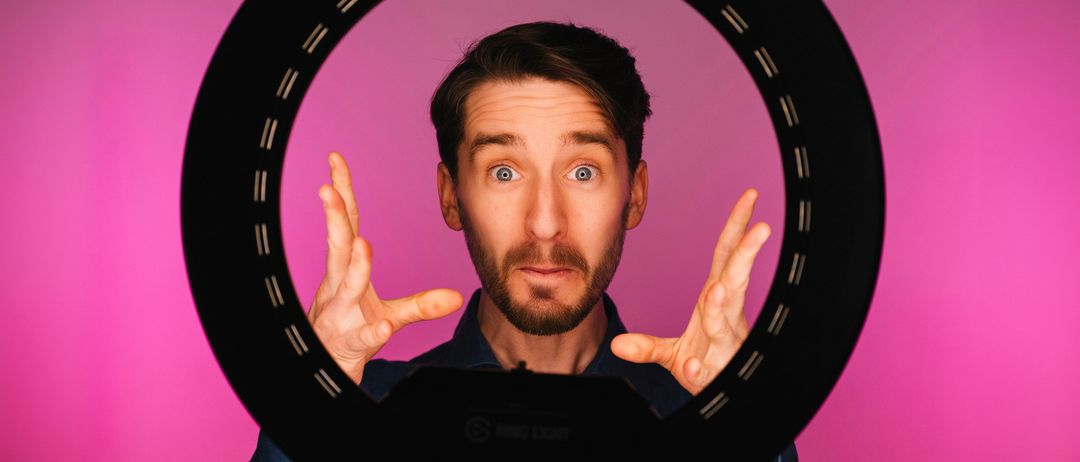 Ein Mann mit Bart posiert mit erhobenen Händen in einem schwarzen kreisförmigen Rahmen vor einem rosa Hintergrund.