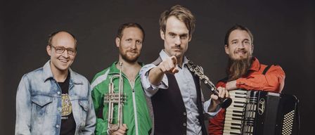 Vier Musiker in lässiger Kleidung stehen zusammen. Einer spielt Trompete, ein anderer Akkordeon, ein Dritter Klarinette und der vierte eine Gitarre. Sie lächeln und posieren für ein Foto.