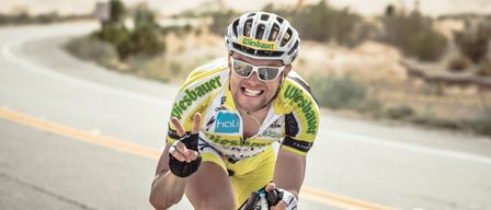 Ein Radfahrer fährt auf einer Straße mit einer Wüstenlandschaft im Hintergrund. Er trägt ein gelbes Trikot, eine Sonnenbrille und einen Helm.