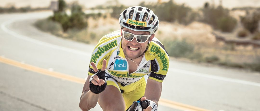Ein Radfahrer fährt auf einer Straße mit einer Wüstenlandschaft im Hintergrund. Er trägt ein gelbes Trikot, eine Sonnenbrille und einen Helm.