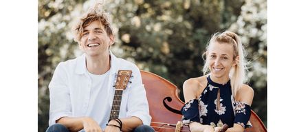 Ein Mann und eine Frau sitzen auf einer Betonkante und lächeln. Der Mann hält eine Gitarre und die Frau eine Flöte. Dahinter steht ein Cello und ein grüner, blattreicher Hintergrund.