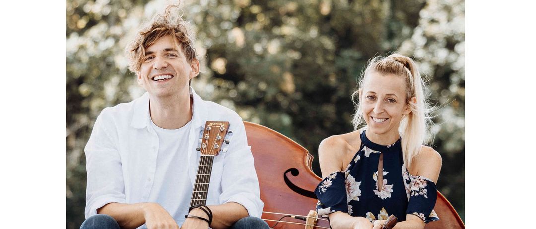 Ein Mann und eine Frau sitzen auf einer Betonkante und lächeln. Der Mann hält eine Gitarre und die Frau eine Flöte. Dahinter steht ein Cello und ein grüner, blattreicher Hintergrund.