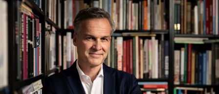 Ein Mann in einem Anzug steht mit verschränkten Armen vor einem Bücherregal voller Bücher und lächelt.