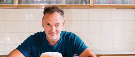 Ein Mann sitzt an einem Küchentresen, lächelt und lehnt sich mit den Ellbogen darauf. Vor ihm steht ein Kuchen auf einem Kuchenständer und eine gelbe Tasse auf einer Untertasse. Dahinter befinden sich Küchenschränke und Regale mit verschiedenen Gegenständen.