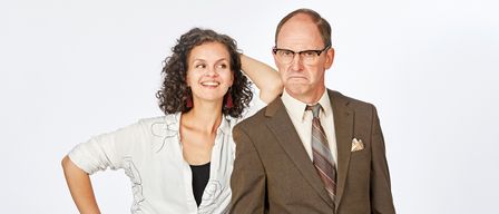 Ein Mann in einem braunen Anzug steht mit geballter Faust, während eine Frau mit lockigem Haar in einem weißen Oberteil und schwarzen Hosen hinter ihm posiert. Sie stehen vor einem schlichten weißen Hintergrund.