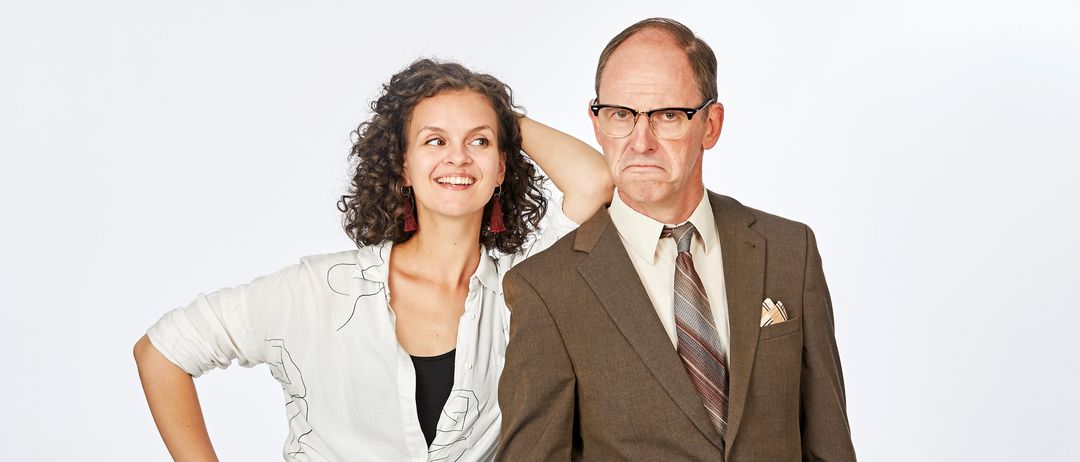 Ein Mann in einem braunen Anzug steht mit geballter Faust, während eine Frau mit lockigem Haar in einem weißen Oberteil und schwarzen Hosen hinter ihm posiert. Sie stehen vor einem schlichten weißen Hintergrund.