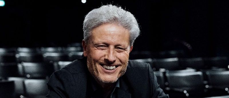 Ein älterer Mann lächelt und sitzt mit verschränkten Armen in einem leeren Theater. Er trägt einen schwarzen Anzug.