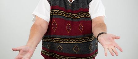 Ein Mann mit grauen Haaren und einer Brille, der eine gemusterte Weste trägt, steht mit offenem Mund und ausgestreckten Armen.