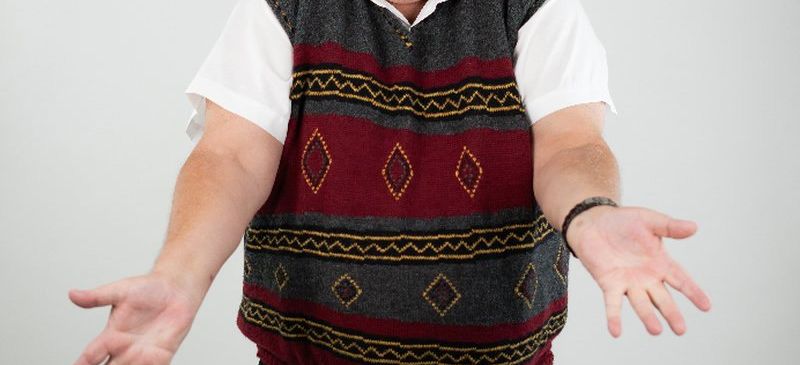 Ein Mann mit grauen Haaren und einer Brille, der eine gemusterte Weste trägt, steht mit offenem Mund und ausgestreckten Armen.