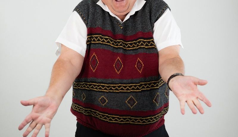 Ein Mann mit grauen Haaren und einer Brille, der eine gemusterte Weste trägt, steht mit offenem Mund und ausgestreckten Armen.