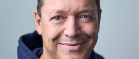 Ein Mann mit grauem Haar, der eine blaue Kapuzenjacke trägt, lächelt in die Kamera. Sein Gesicht zeigt Falten und einen warmen Ausdruck.