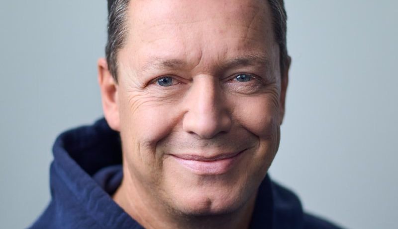 Ein Mann mit grauem Haar, der eine blaue Kapuzenjacke trägt, lächelt in die Kamera. Sein Gesicht zeigt Falten und einen warmen Ausdruck.
