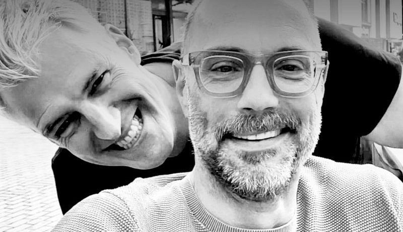 Zwei Männer lächeln auf einem Schwarz-Weiß-Foto, einer mit Brille und Bart, der andere mit schelmischem Ausdruck.