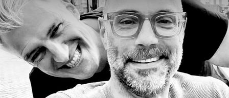Zwei Männer lächeln auf einem Schwarz-Weiß-Foto, einer mit Brille und Bart, der andere mit schelmischem Ausdruck.
