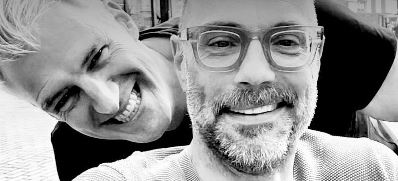 Zwei Männer lächeln auf einem Schwarz-Weiß-Foto, einer mit Brille und Bart, der andere mit schelmischem Ausdruck.