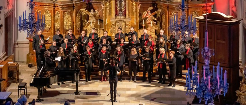 Ein Chor in Schwarz und Rot singt auf einer Bühne in einer großen Kirche, mit einem Publikum, das unten sitzt.