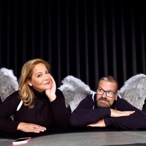Eine Frau und ein Mann in Engelsflügeln sitzen an einem Tisch und posieren für ein Foto. Die Frau lächelt, während der Mann die Arme verschränkt hat. Hinter ihnen befindet sich ein schwarzer Vorhang.