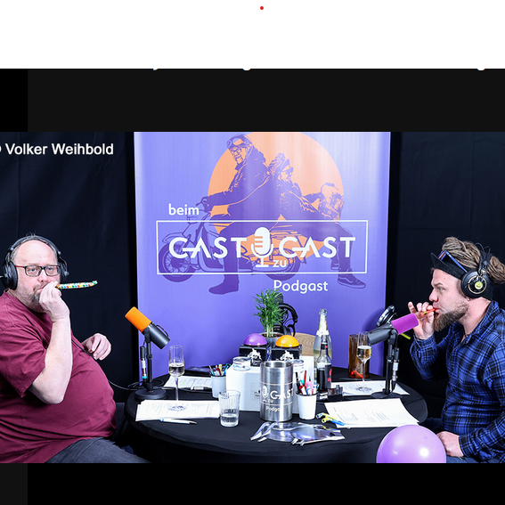 Two men sit on either side of a table with microphones. One is smoking a pipe and the other is drinking from a glass. Behind them is a banner with the logo of CASTCAST Podcast.