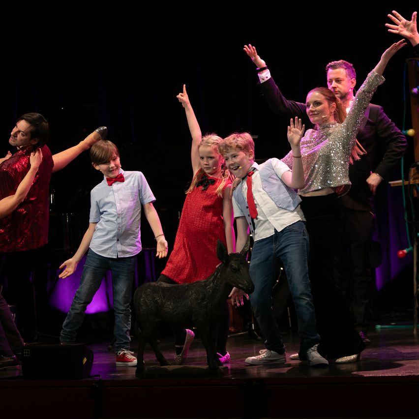 Eine Gruppe von Menschen, darunter Erwachsene und Kinder, tritt auf einer Bühne auf. Sie heben die Hände und lächeln, vor ihnen steht ein kleiner schwarzer Ziegenbock.