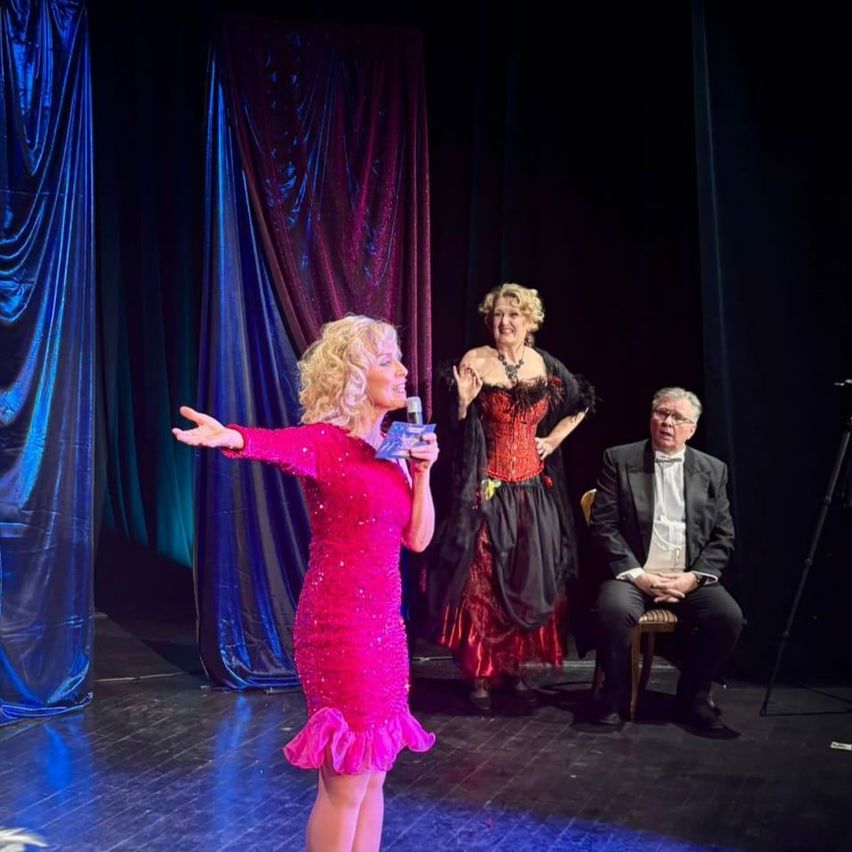 Eine Frau in einem rosa Kleid steht auf der Bühne und hält ein Mikrofon, während ein Mann in einem Anzug auf einem Stuhl sitzt und eine andere Frau in einem roten Kleid hinter ihr steht.