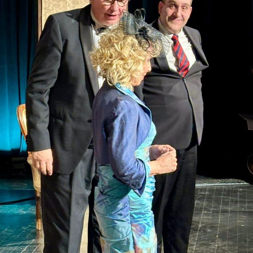 Drei Personen stehen auf einer Bühne, zwei Männer in Anzügen und eine Frau in einem blauen Kleid. Die Frau trägt einen schwarzen Hut und hält ihre Hände zusammen. Die Männer haben Lächeln auf ihren Gesichtern.