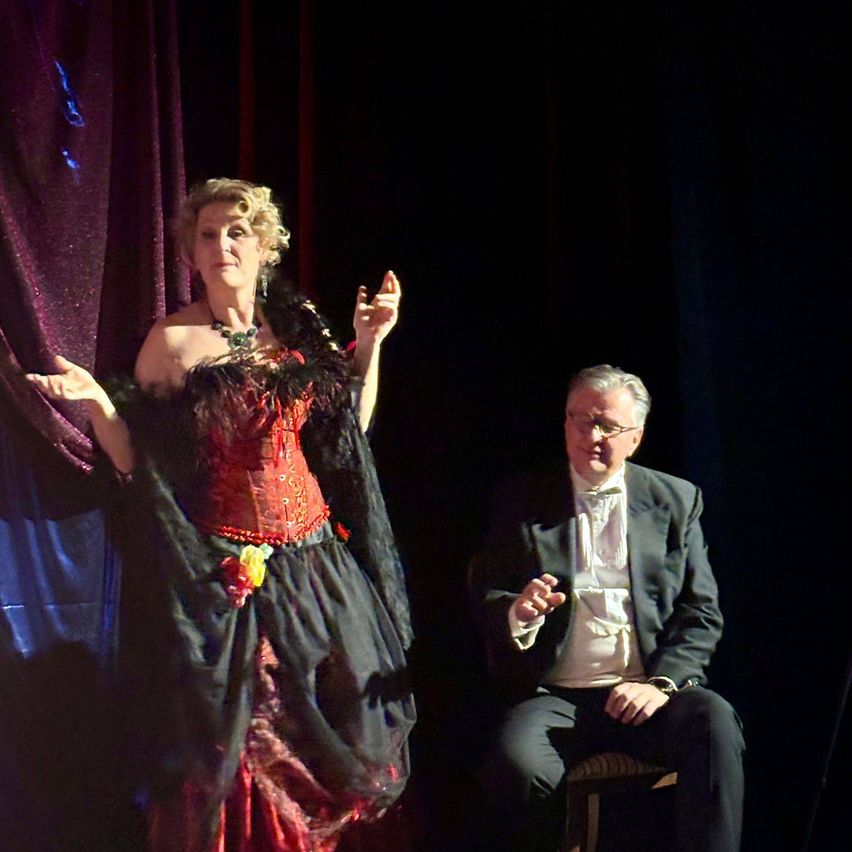 Eine Frau in einem roten Korsett und schwarzem Kleid, mit einer gelben Blume, gestikuliert auf der Bühne. Neben ihr sitzt ein Mann in einem schwarzen Anzug und Fliege auf einem Stuhl und schaut zu.