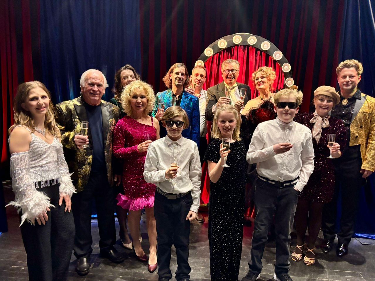 Eine Gruppe von Menschen steht zusammen auf einer Bühne, einige in Kostümen gekleidet, halten Champagnergläser in der Hand und lächeln. Sie scheinen ein Ereignis mit einem roten Vorhang im Hintergrund zu feiern.