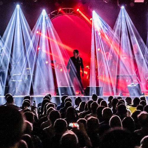 A live concert with a performer on stage, surrounded by red and white lights. The audience is watching, some are taking photos.