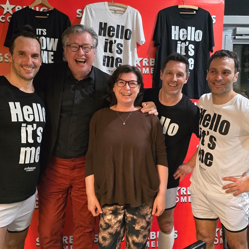 Fünf Personen stehen vor einer roten Wand, alle tragen verschiedene T-Shirts mit dem Slogan 'Hallo, ich bin's'. Zwei Männer tragen schwarze Shirts, einer weiß und die Frau braun. Sie alle lächeln.