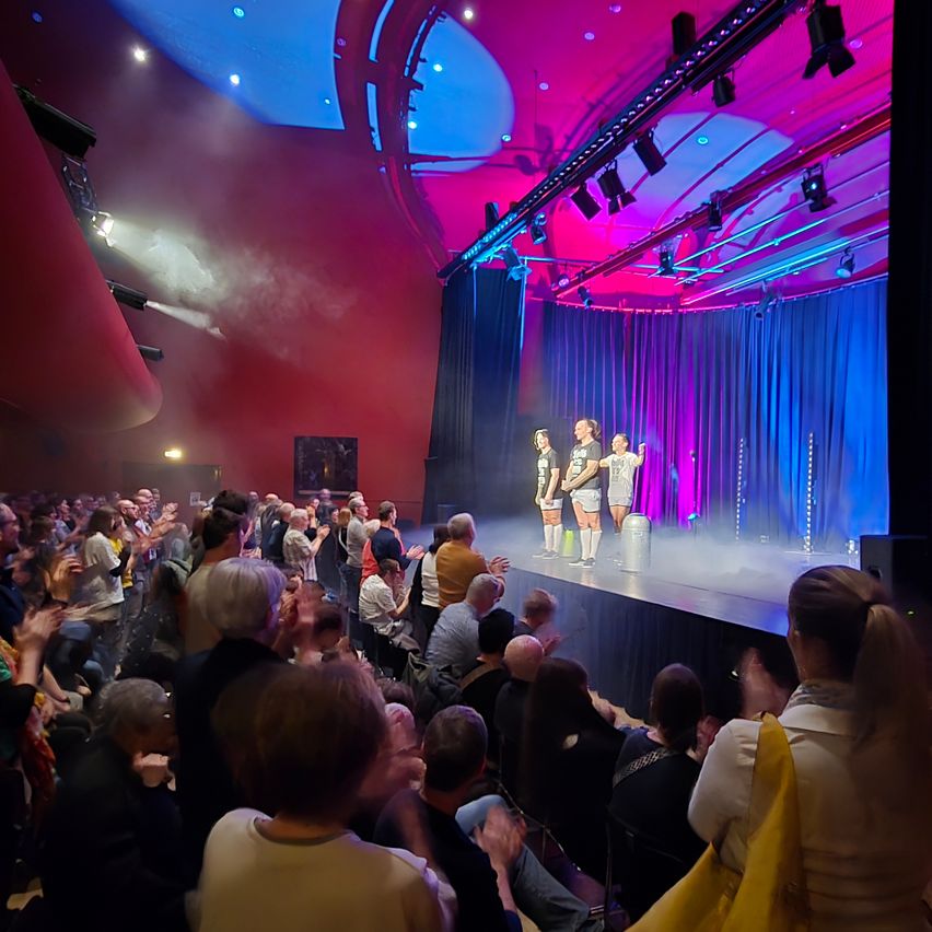Eine Bühne mit drei Darstellern vor einem Publikum. Die Bühne ist mit blauen und rosa Lichtern beleuchtet, und das Publikum klatscht.