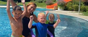 Eine Frau und drei Kinder lächeln und spielen in einem Schwimmbad mit Wasserrutsche und Pflanzen im Hintergrund.