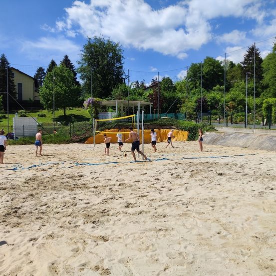 Eine Gruppe von Menschen spielt Volleyball auf einem sandigen Spielfeld, umgeben von Bäumen und einem Haus. Der Himmel ist blau mit einigen Wolken.