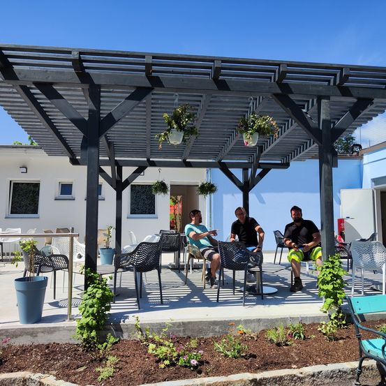 Drei Männer sitzen unter einer hölzernen Pergola auf einer Terrasse mit mehreren Stühlen und Topfpflanzen. Dahinter steht ein weißes Gebäude mit blauer Umrandung.