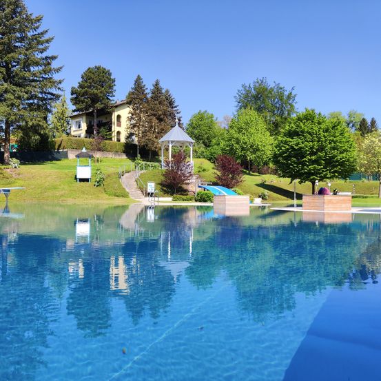 Ein großer Swimmingpool ist von üppiger Grünfläche und Bäumen umgeben. Im Hintergrund steht ein gelbes Haus mit mehreren Fenstern auf einem Hügel. Ein Pavillon und eine Bank befinden sich in der Nähe des Pools.