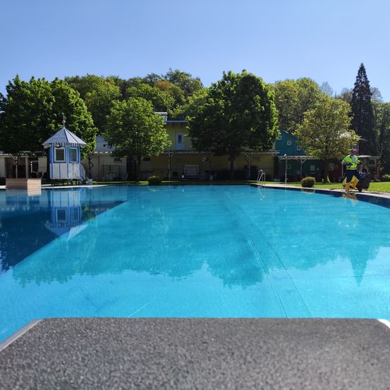 Ein großer Swimmingpool mit blauer Wasserfläche ist von einem grünen Rasen umgeben. Ein kleines blaues und weißes Gebäude steht in der Ecke. Eine Person in einem gelben Vest steht in der Nähe des Pools. Bäume und Gebäude sind im Hintergrund.
