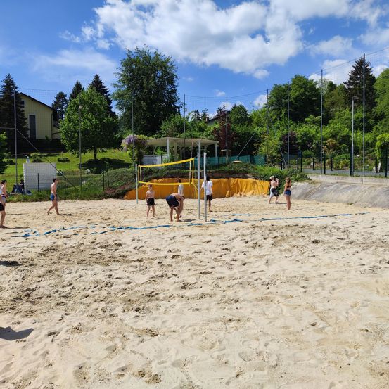 Eine Gruppe von Menschen spielt Beachvolleyball auf einem sandigen Platz mit einem gelben Netz. Einige stehen, während andere sich beugen. Bäume und Büsche sind im Hintergrund.