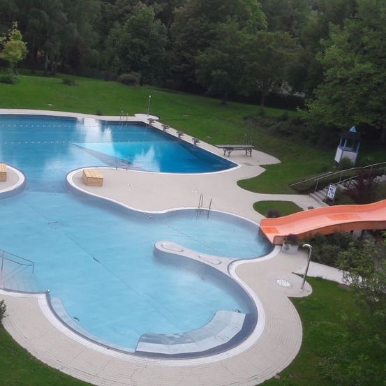 Eine Luftaufnahme eines Schwimmbeckens mit Wasserrutsche, Holzbänken und einem Rasenbereich mit Bäumen.