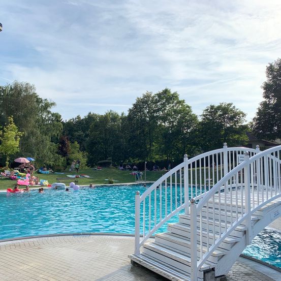 Ein sonniger Tag an einem Freibad mit einem weißen Handlauf und mehreren Schwimmenden, einige unter Sonnenschirmen.