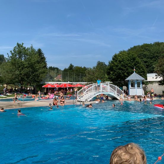 Ein Schwimmbad mit einer Brücke und Menschen, die schwimmen und sich entspannen. Im Hintergrund sind Bäume und ein Gebäude zu sehen.