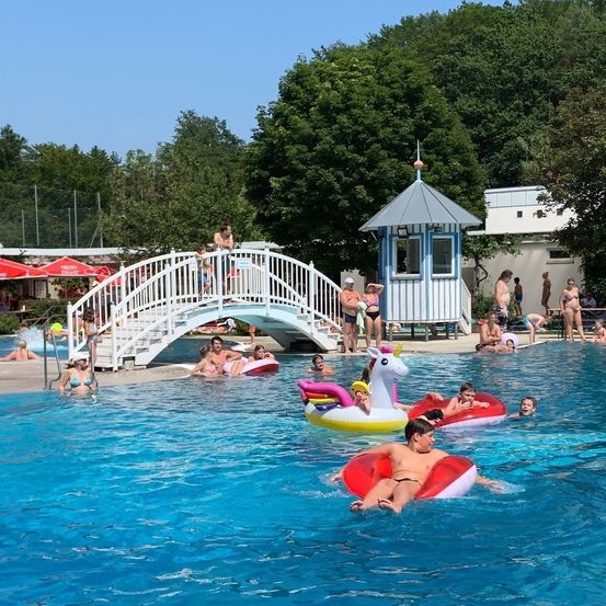 Eine Gruppe von Menschen schwimmt in einem Pool. Einige treiben auf aufblasbaren Spielzeugen. Eine weiße Brücke führt zu einer Umkleidekabine. Bäume und ein Gebäude sind im Hintergrund.