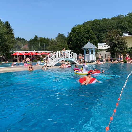 Ein großer Außenpool voller Menschen, die das Wasser genießen, einige in aufblasbaren Schwimmhilfen. Eine weiße Brücke verbindet den Poolbereich mit einem Gebäude mit rotem Dach. Bäume und ein klarer Himmel sind im Hintergrund zu sehen.