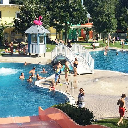 Ein geschäftiger Außenpoolbereich mit schwimmenden Menschen, einer Rutsche und einem Rettungsschwimmerturm. Bäume und Häuser umgeben den Bereich.